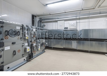 The setting features a large ventilation system installed in an industrial building. The area is bright with metal equipment arranged along the walls and pipes above.