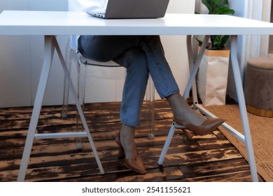 The setting shows a woman engaged in work on her laptop at a contemporary desk. Her relaxed posture and chic footwear complement the modern decor of the space. - Powered by Shutterstock