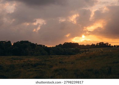 Setting sun glowing through dramatic clouds over a peaceful rural landscape with hills, a field, and trees