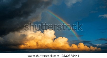 The setting sun illuminates a dark storm cloud on the left and golden-orange cumulus clouds. A vibrant rainbow arcs across the blue sky, creating a stunning scene.