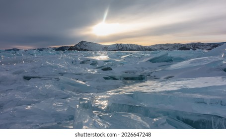 The setting sun illuminates the ice hummocks lying in disarray on the frozen lake. Highlights on shiny edges. A mountain range against the background of a cloudy evening sky. Baikal