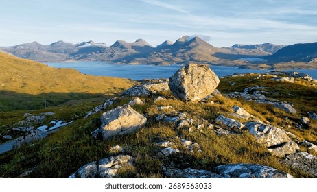 The setting sun illuminates an unstable boulder on hill, overlooking the mountain range and Loch Torridon, Scotland.