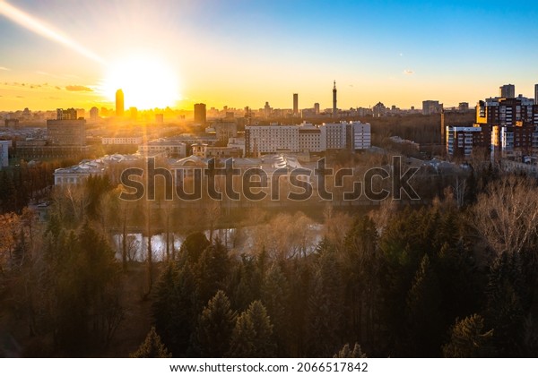 setting sun over the city and\
the city park, rays over skyscrapers and fir trees in the\
foreground.