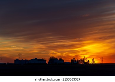 A setting sun reflects gold and yellow on clouds and silhouettes farm buildings on the horizon.