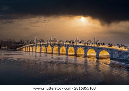 The setting sun shines through the clouds at the Summer Palace's Seventeen-Arch Bridge, casting golden light through the arches onto the frozen lake, creating a magnificent scene.