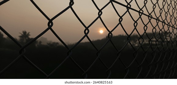 A setting sun is visible through a chain-link fence, silhouetting the surrounding landscape.  The hazy atmosphere adds a serene, slightly melancholic mood.
