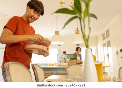 Setting table with plates, smiling asian teenage man in kitchen with family in background. dining, preparation, happiness, togetherness, domestic, lifestyle - Powered by Shutterstock