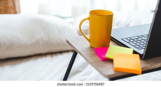 Setup of laptop on portable table with coffee and tea mug and sticky notes on bed with stack of documents and papers with graphs and charts placed on clean white sheet on bed in room at home