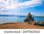 A Sevan Monastery Sevanavank with a steeple sits on a hill overlooking a body of water. The sky is clear and the water is calm
