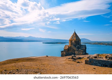 A Sevan Monastery Sevanavank with a steeple sits on a hill overlooking a body of water. The sky is clear and the water is calm