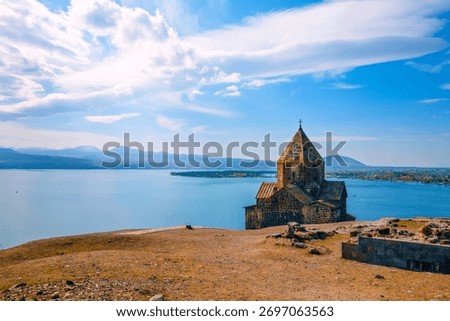 A Sevan Monastery Sevanavank with a steeple sits on a hill overlooking a body of water. The sky is clear and the water is calm