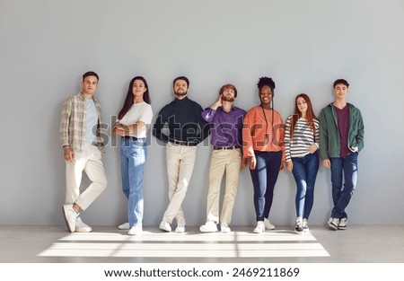 Seven confident people of different ethnicities are standing facing forward with relaxed posture. Stylish smiling young men and women stand in row against background of wall in room with sunlight.