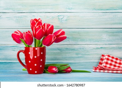 Seven red tulips bouquet in a red glass, The floor has two red tulips and one handkerchief placed in front of the blue wooden wall.