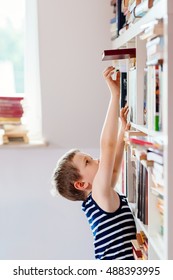 Seven Years Old Boy Reaching For Book In Library. Child Reading