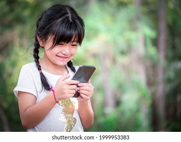 A seven-year-old Asian girl is smiling happily as she uses her mobile phone to search for information while on a family trip to the park.