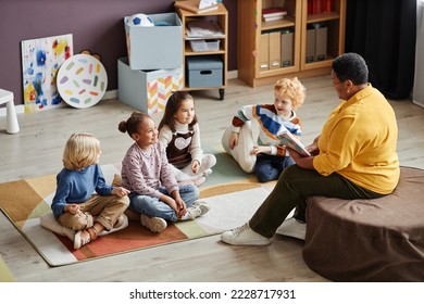 Several adorable intercultural kids gathered on the floor in front of their teacher of nursery school reading them book of tales - Powered by Shutterstock