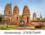 Several ancient, weathered red-brick prangs (towers) at a historical site in Ayutthaya, Thailand. In the foreground, the remains of low walls and stone structures are visible under a clear, blue sky.