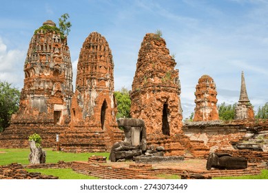 Several ancient, weathered red-brick prangs (towers) at a historical site in Ayutthaya, Thailand. In the foreground, the remains of low walls and stone structures are visible under a clear, blue sky.