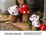 Several brightly painted figurine of a fly agaric, featuring red and white polka dots, are set amidst garden pathways, surrounding area includes rocks, gravel and scattered greenery. Garden decoration