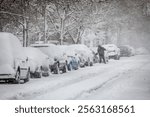 Several Cars parked in an urban Street covered in heavy Snow, one Man brushing off Snow from his Car; Copy Space