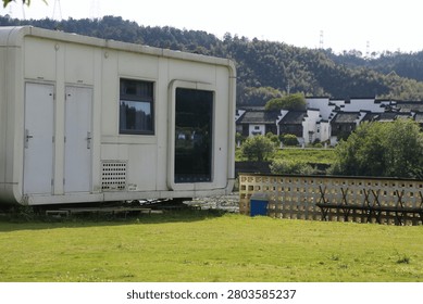 Several compact white cabin structures stand along a grassy area near a low fence. The background includes a riverbank wall, distant buildings, wooded hills, and bright natural light across the open lawn.