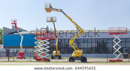 Several different wheeled scissor lifts and wheeled articulated lifts with telescoping boom and basket on an asphalt ground against the sky and an industrial building
