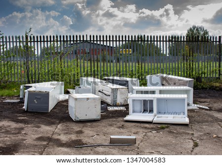 Several discarded fly-tipped fridges and freezers dumped on concrete ground in front of spiked security fence, Clayton, Manchester, UK.