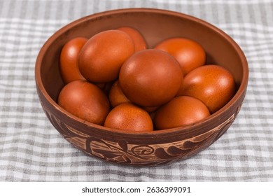 Several Easter eggs painted brown in a clay bowl decorated with plant pattern on a table covered with checkered tablecloth