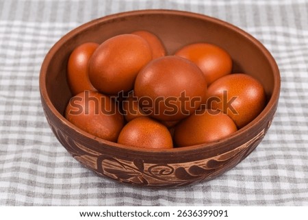 Several Easter eggs painted brown in a clay bowl decorated with plant pattern on a table covered with checkered tablecloth