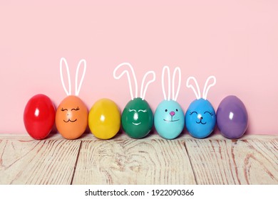 Several eggs with drawn faces and ears as Easter bunnies among others on white wooden table against pink background