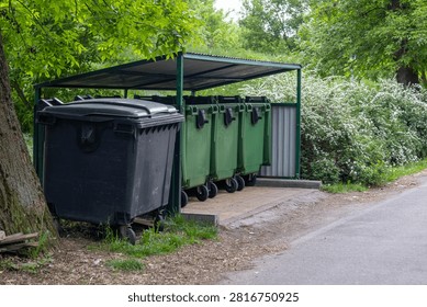 Several garbage bins are placed under a shelter next to a road. Trees and bushes surround the area, showing a typical urban setting.