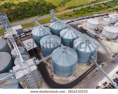 Several large grain silos are shown from above in an industrial area surrounded by fields. Transport tracks are visible nearby, indicating movement of goods.