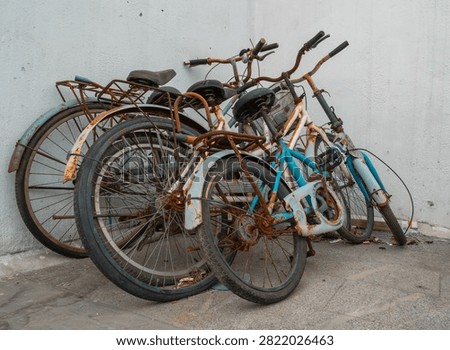 Several old bicycles with rusted frames and worn tires are piled near a white wall, showing signs of neglect