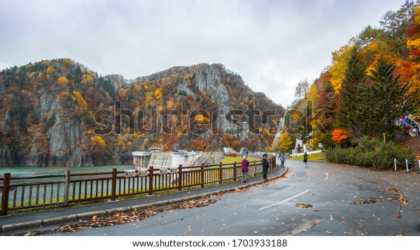 Several people are\
standing on a road near the lake in an autumn season surrounded by\
yellow and orange leaves tree. There\'s a damn and large cliffs in\
the background.