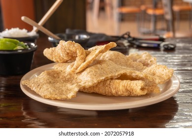 Several Pieces Of Pork Rinds Served On A Beige Plate, The Background Is Blurred And You Can See Several Black Containers Containing Split Lemons And Chopped Onion