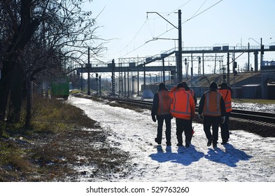 Several Railway Workers In Signaling Dirty Orange Uniforms Are On The Road Next To The Railway Line. The Train Crew Goes To Work In The Winter Sunny Morning.