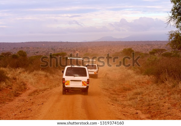 Several safari cars in Kenya in Africa, red\
sand and mountains in the\
background