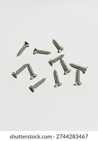 Several small silver Phillips-head screws are scattered randomly across a smooth white surface, each showing sharp pointed tips, deep spiral threads, and reflective metallic finishes under soft lighting.