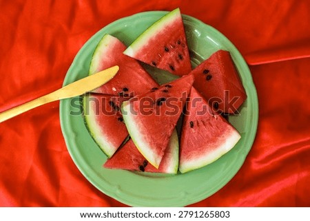 Several triangular watermelon slices are arranged on a green ceramic plate with a yellow wooden knife laid across them, the top-down watermelon composition pops boldly against the crumpled red fabric tablecloth below, and this overhead watermelon flat lay delivers a vibrant and graphic summer food aesthetic