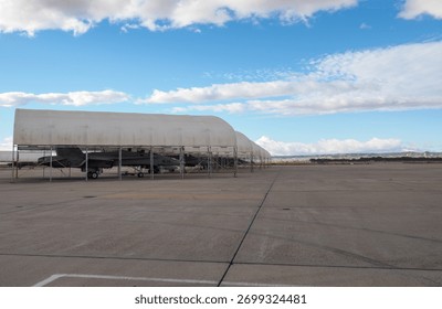 Several twin-engine multirole fighter jets parked in open shelters on an airbase apron under a clear sky