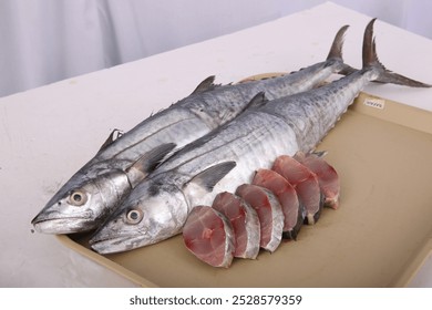 Several whole kingfish lie on a tray, their silvery scales shimmering in the light. A few pieces of kingfish fillets are arranged below, showcasing the fish's firm white flesh.