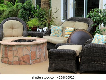 Several Wicker Chairs And Couch Surrounding Beautiful Stone Fire Pit In Backyard Garden.