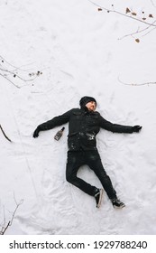 A Severely Drunk Homeless Man In A Black Coat And Hat Lies, Asleep On His Back On White Snow In The Cold Winter With A Bottle Of Alcohol. Photography, Concept.