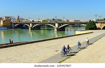 Seville, Spain - Feb 17, 2016: Family Bike Ride Along The Guadalquivir River Bank With The Bridge And Triana Neighborhood In The Background