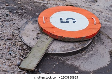 The Sewer Manhole At The Construction Site Is Covered With A Round Orange Sign With The Number Five. Designation Of The Object At Work On The Reconstruction Of Water Supply And Sanitation Systems.