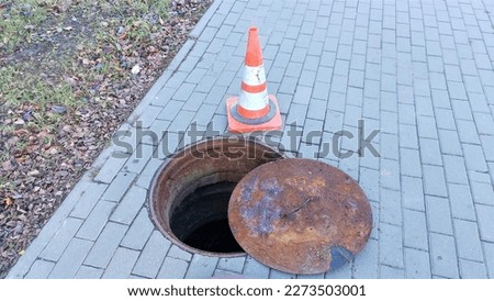 The sewer manhole is open on the paved concrete sidewalk near the grassy lawn. Next to it lies an iron lid and a brightly painted plastic bollard for safety