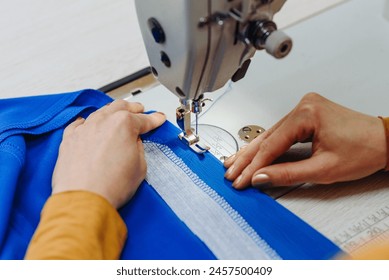sewing machine with needle and thread on table in atelier, close-up view of hands of working needlewoman, blue fabric and needle, handmade and sewing concept
