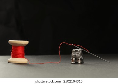 Sewing needle with red thread and thimble on grey table against black background