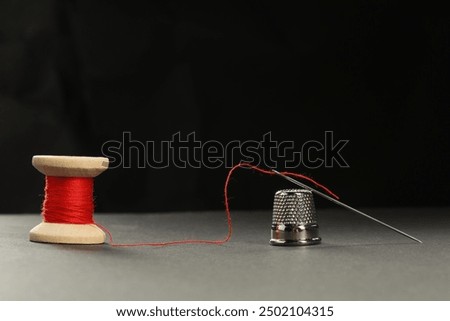 Sewing needle with red thread and thimble on grey table against black background