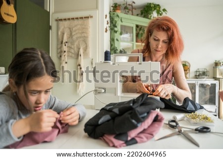 Sewing process. Female stitching fabric on professional manufacturing machine at home while her little daughter helping to her with dress production 